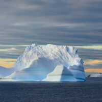Türkis gestreifter Eisberg im Licht der gerade aufgehenden Sonne, im Hintergrund aprikotfarben der Himmel mit Tafeleisbergen vor der Landzunge einer Insel im Archipel der Antarktischen Halbinsel.