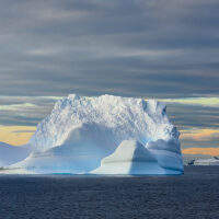 Türkis gestreifter Eisberg im Licht der gerade aufgehenden Sonne, im Hintergrund aprikotfarben der Himmel mit Tafeleisbergen vor der Landzunge einer Insel im Archipel der Antarktischen Halbinsel.