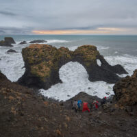 Fünf Fotografen bei ihren Aufnahmen des Gatklettur Felsentors in schäumender Brandung an der Basaltküste bei Arnarstapi vor einer Wetterfront am Horizont mit Morgenrot.