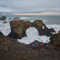 Fünf Fotografen bei ihren Aufnahmen des Gatklettur Felsentors in schäumender Brandung an der Basaltküste bei Arnarstapi vor einer Wetterfront am Horizont mit Morgenrot.