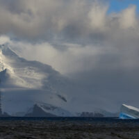 A snow-covered mountain peak on Coronation Island glows in the light of a sunbeam piercing the dense, low-hanging cloud cover, while a small, tilted iceberg floats past in the foreground.